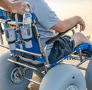 Wheeleez Sandcruiser All-Terrain Beach Wheelchair Rear View In Mediterranean Blue Color With Large Balloon Wheels And RoboCup Holder, Positioned On A Sandy Beach With Man Sitting In The Wheelchair While A Man Stands Behind The Wheelchair Holding The Handles