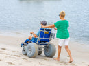 Wheeleez Sandcruiser All-Terrain Beach Wheelchair Rear View In Mediterranean Blue Color With Large Balloon Wheels And RoboCup Holder, Positioned On A Sandy Beach With Man Sitting In The Wheelchair While A Woman Pushes The Wheelchair And Ocean In The Background.