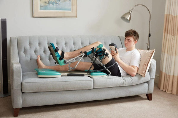 A Man Using The Kinetec Spectra Essential Knee CPM Machine Which Is Used For Knee Rehabilitation On A Sofa
