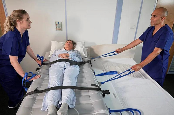 A Patient In A Light Blue Hospital Gown Lies On A Gurney Bed While Two Healthcare Workers In Blue Scrubs Use Blue Straps To Transfer The Patient, Using The Winncare Mangar Swift. The Scene Is Set In A Clinical Room With White Walls And Medical Equipment