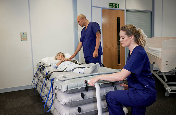 A Patient Lies On The Winncare Mangar Rhino With Airflo Duo, While Two Healthcare Workers In Blue Scrubs Attend To Her In A Hospital Room