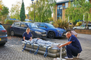 A Person In A Hospital Parking Lot Using The Winncare Mangar Rhino With Airflo Duo With Two Healthcare Workers In Blue Scrubs Nearby, With Parked Cars And A Building In The Background 