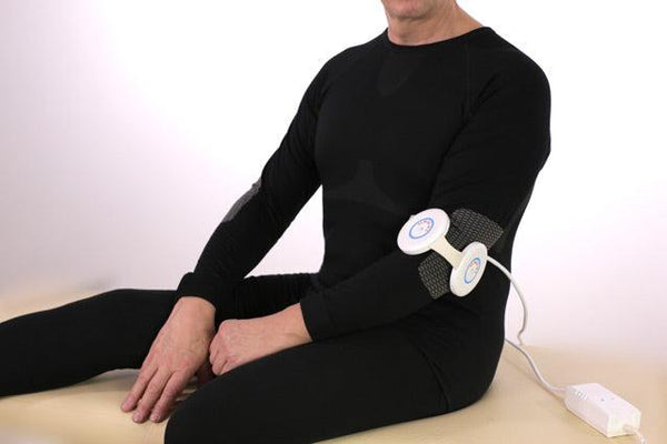 A Person Wearing A Black Long-Sleeve Compression Garment Sits Cross-Legged On A Beige Surface Against A White Background, With The Almagia International ALMAG-01 PEMF Therapy Device Attached To The Arm And A White Power Cord Visible