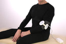 A Person Wearing A Black Long-Sleeve Compression Garment Sits Cross-Legged On A Beige Surface Against A White Background, With The Almagia International ALMAG-01 PEMF Therapy Device Attached To The Arm And A White Power Cord Visible