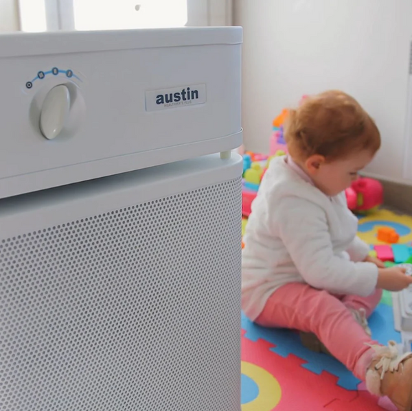 A Sandstone Austin Air Systems HealthMate Plus Standard Air Purifier In The Foreground, With A Baby Sitting On A Colorful Play Mat In The Background
