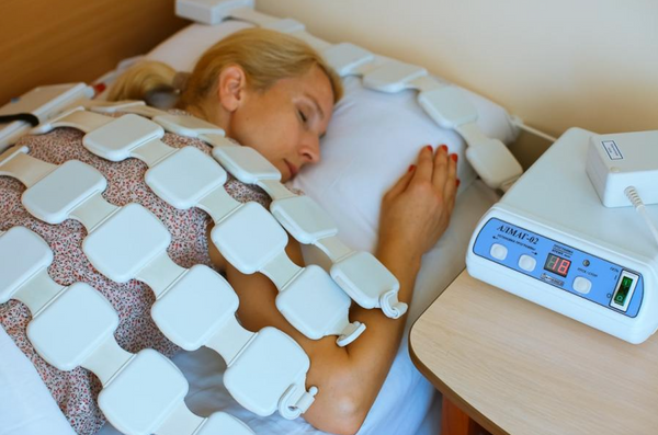A Woman Lying Down On A Treatment Bed Wearing An ALMAG-2 White Electrode Pad Device On Her Body, With A Control Unit On A Bedside Table Nearby
