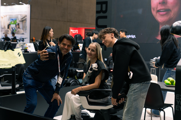 A Woman With A Robotic Arm Is Sitting In The STRUTT ev1 Smart Electric Mobility Vehicle While A Man Standing Next To Her Is Taking A Selfie Of Her Himself And A Young Man Standing On The Other Side Of Her In A Convention Center Setting