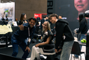 A Woman With A Robotic Arm Is Sitting In The STRUTT ev1 Smart Electric Mobility Vehicle While A Man Standing Next To Her Is Taking A Selfie Of Her Himself And A Young Man Standing On The Other Side Of Her In A Convention Center Setting