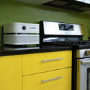 Austin Air Systems "it" Personal Compact Air Purifier Placed On A Black Countertop Next To A Stainless Steel Gas Stove In A Kitchen With Bright Yellow Cabinets And A Green Wall