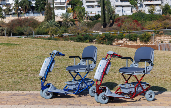Centered View Two Tzora Feather Ultra Lightweight Mobility Scooters Shown In Red And Blue Colors During The Day With Buildings Seen In The Background