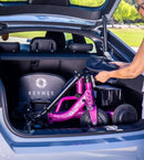 Close Up Rear View Of An Elderly Woman Loading The Reyhee Triad Compact Folding Electric Mobility Scooter In The Violet Frame Color In The Trunk Of Her Compact Car In A Daytime Outdoor Setting