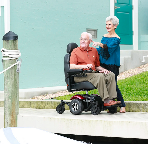 Vista de cerca lateral derecha de un hombre mayor conduciendo la silla de ruedas eléctrica Merits Vision Sport en color rojo en un muelle, con una mujer mayor de pie a su lado señalando algo a lo lejos