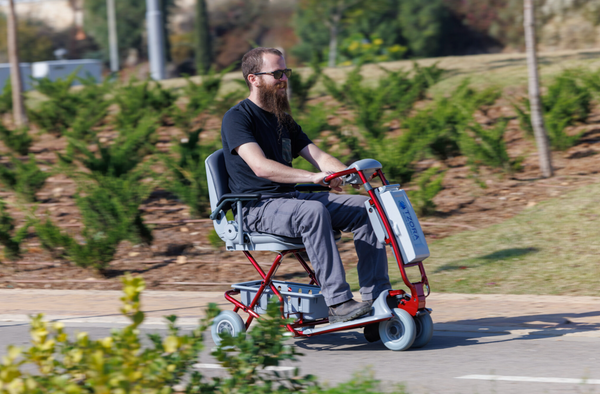 Close Up View Of A Man Riding The Tzora Feather Ultra Lightweight Mobility Scooter Shown In The Red Color During The Day