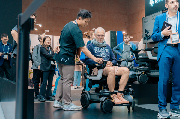 Close Up View Of An Elderly Man Sitting In The STRUTT ev1 Smart Electric Mobility Vehicle While Another Man Is Standing Next To Him Explaining The Mobility Device In A Convention Center Setting