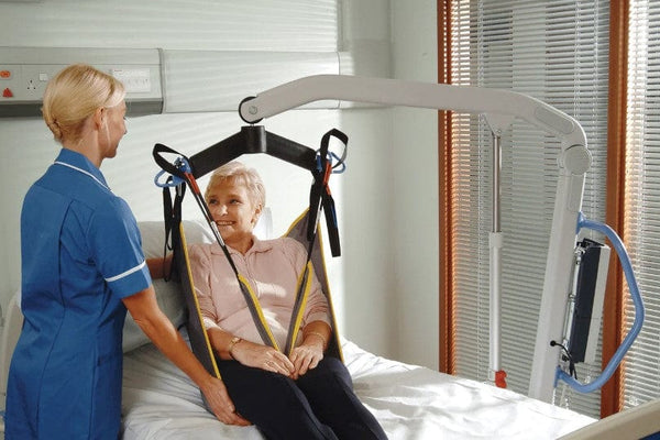 Close Up View Of An Elderly Woman In A Sling Being Carried By The Joerns Hoyer® Presence Pro HD Patient Lift With A Healthcare Worker Assisting Her In A Hospital Room Setting