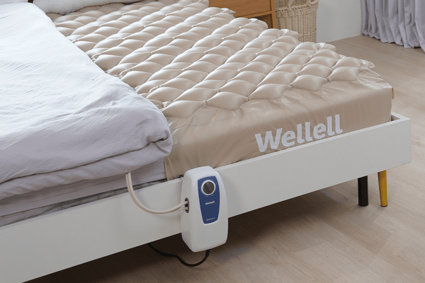 Close Up View Of The Wellell Domus 1 Alternating Pressure Redistribution Mattress Shown On A Platform Bed Frame With The Control Unit Hanging Off The Side Of The Bed Frame In A Home Care Setting