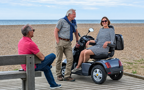 Close Up View Of Three People Conversing With The Ocean In The Background Shown With A Woman Sitting On The AFIKIM Afiscooter S4 Scooter In The Dark Gray Color In The Turned Position