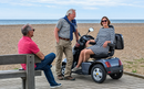 Close Up View Of Three People Conversing With The Ocean In The Background Shown With A Woman Sitting On The AFIKIM Afiscooter S4 Scooter In The Dark Gray Color In The Turned Position