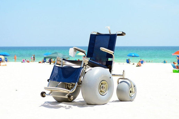Debug Mobility All-Terrain Beach Wheelchair Shown On A Sandy Beach With The Ocean In The Background. The Wheelchair Has Large Durable-Looking Wheels And Is Designed For Accessibility And Mobility On Various Terrains. 