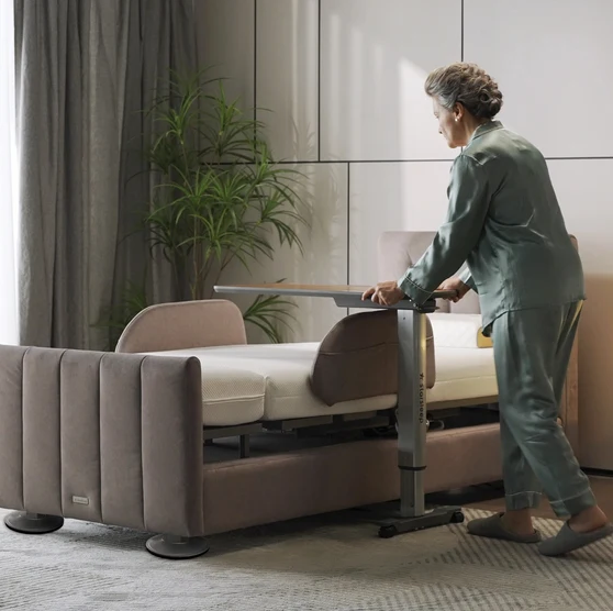 Elderly Woman Moving The StarSleep Overbed Table Under The StarSleep Arlo Rotating Bed Shown In A Bedroom Setting