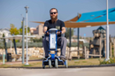 Front Centered View Of A Man Riding The Tzora Feather Ultra Lightweight Mobility Scooter Shown In The Blue Color During The Day In A Park Setting