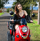 Front Centered View Of The Pushpak Motors- Pushpak 1000 Two- Person Electric Scooter In The Red Color Shown With A Woman Riding In The Countryside