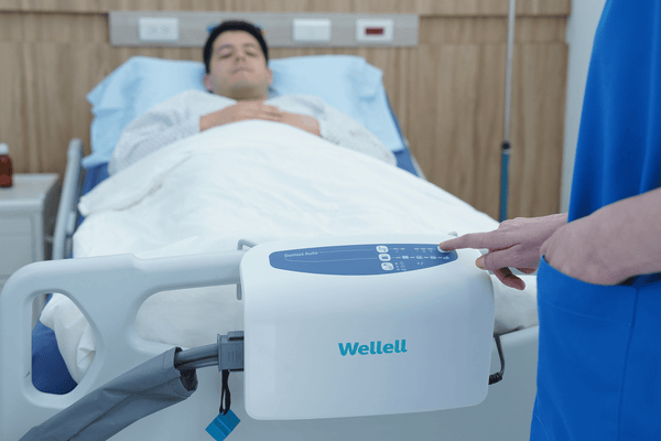Front Centered View Of The Wellell Domus Auto Pressure Redistribution Mattress System With Quilted Cover Shown On An Adjustable Hospital Bed In A Hospital Room With Patient Lying On Mattress With Healthcare Worker Adjusting The Settings On The Control Unit Hanging On The Footboard Panel