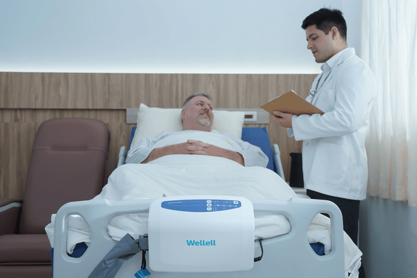 Front Centered View Of The Wellell Domus Auto Pressure Redistribution Mattress System With Quilted Cover Shown On An Adjustable Hospital Bed In A Hospital Room With Patient Lying On Mattress With Healthcare Worker Conversing With Him