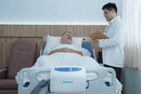 Front Centered View Of The Wellell Domus Auto Pressure Redistribution Mattress System With Quilted Cover Shown On An Adjustable Hospital Bed In A Hospital Room With Patient Lying On Mattress With Healthcare Worker Conversing With Him
