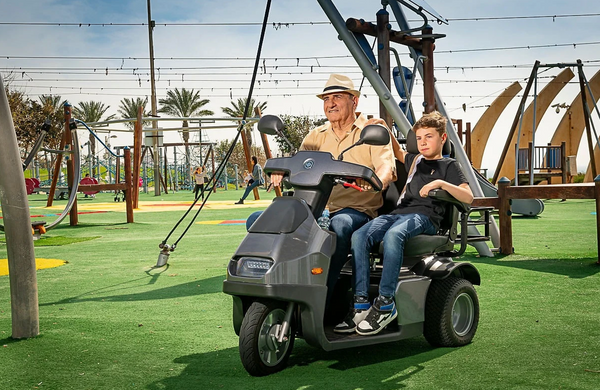 Front Left Side View Of The AFIKIM Afiscooter S3 Scooter In Dark Gray Color With Optional Dual Seat Configuration Shown With An Elderly Man And Young Boy Riding The Scooter In A Playground Setting