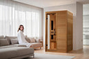 Front Left Side View Of The Dynamic Lucca 2 Person Ultra Low EMF FAR Infrared Indoor Sauna | DYN-6215-05 Elite Shown With A Woman Sitting On A Couch Next To The Sauna In An Indoor Setting