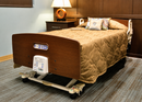 Front Left Side View Of The Joerns DolphinCare™ Integrated Fluid Immersion Simulation Complete Bed System Shown In An Indoor Setting On A Wooden Motorized Hospital-Style Bed Frame With A Quilted Tan Bedspread, Shown In A Bedroom Setting With A Nightstand, Lamp, And Carpeted Floor