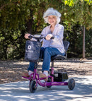 Front Left Side View Of The Reyhee Triad Compact Folding Electric Mobility Scooter With The Violet Colored Frame Shown In A Sunny Day Setting With An Elderly Woman Riding The Scooter