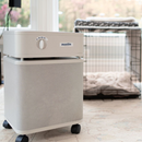 Front Right Side View Of The Austin Air Systems HealthMate Standard Air Purifier Shown In The Sandstone Color, Branded 'austin', Shown In A Home Setting With A Dog Crate In The Background