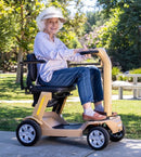 Front Right Side View Of The Reyhee Flex Electric Folding Mobility Scooter With A Tan Frame In An Outdoor Setting During The Day With An Elderly Woman Driving