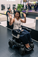 Front Right Side View Of The STRUTT ev1 Smart Electric Mobility Vehicle Shown With A Smiling Woman Sitting In The Chair In A Convention Center Setting