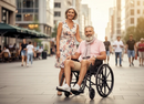 Front View Of An Elderly Man Sitting In The Strongback ErgoSteel Wheelchair With An Elderly Woman Standing Beside Him In A City Setting During The Day