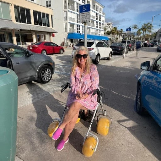 Front View Of The AccessRec AccessWalk® All-Terrain Beach Rollator Shown In The Grey And Black Color With Elderly Woman Sitting In The Chair On A Sunny Day In The City