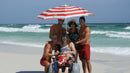 Front View Of The Debug Mobility All-Terrain Beach Wheelchair Shown With A Group Of People Standing Around The Wheelchair Under An Umbrella In A Beach Setting With The Ocean In The Background