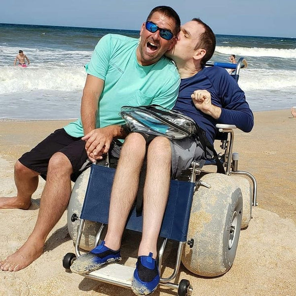 Front View Of The Debug Mobility All-Terrain Beach Wheelchair Shown With One Man Sitting In The Wheelchair With Another Man Leaning Beside Him In A Beach Ocean Setting