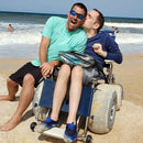 Front View Of The Debug Mobility All-Terrain Beach Wheelchair Shown With One Man Sitting In The Wheelchair With Another Man Leaning Beside Him In A Beach Ocean Setting