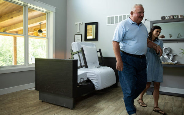 Front View Of The Med Mizer Active Care Deluxe Rotating Bed And Bed In The Chair Position In A Home Setting With An Elderly Man And Young Woman Walking In The Foreground