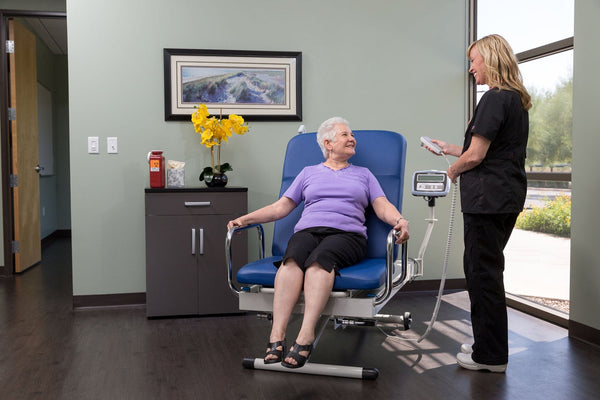 Front View Of The Med Mizer UpScale Adjustable Treatment Table With Scale Elderly Woman Sitting While Clinical Technician Uses Remote Control