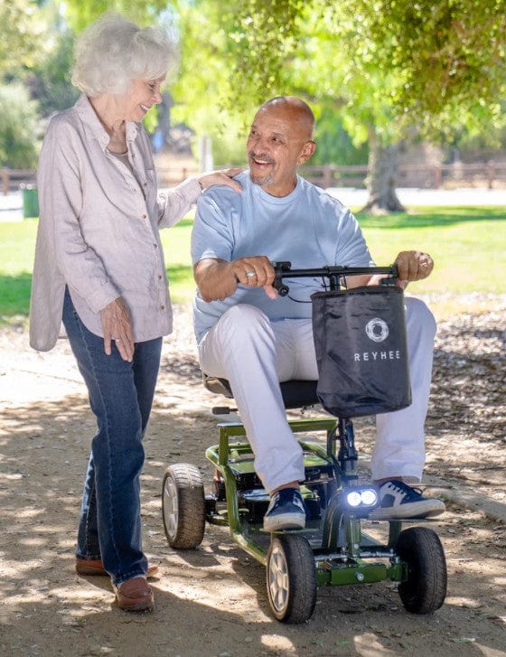 Front View Of The Reyhee EXO Electric Mobility Scooter In Green Color In A Park Setting During The Day With An Elderly Man Sitting In The Chair With An Elderly Woman Standing Next To Him