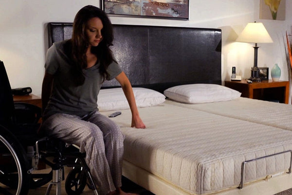 Front View Of The Transfer Master Supernal Hi Low Adjustable Bed Split King Size With A Woman In A Wheelchair Transferring Onto Bed