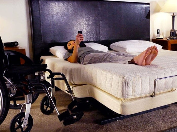 Front View Of The Transfer Master Supernal Hi Low Adjustable Bed With Woman Lying Down Using The Remote Control With A Wheelchair At The Side