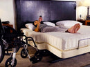 Front View Of The Transfer Master Supernal Hi Low Adjustable Bed With Woman Lying Down Using The Remote Control With A Wheelchair At The Side