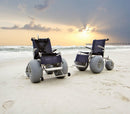 Front View Of Two Models Of The Debug Mobility All-Terrain Beach Wheelchair Shown In A Beach Setting With The Sun In The Background
