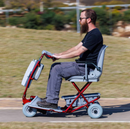 Left Side View Of A Person Riding The Tzora Feather Ultra Lightweight Mobility Scooter Shown In The Red Color Up A Hill On A Sunny Day