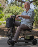 Left Side View Of An Elderly Man Riding The Reyhee Triad Compact Folding Electric Mobility Scooter With The Gun Metal Frame In An Outdoor Setting During The Day